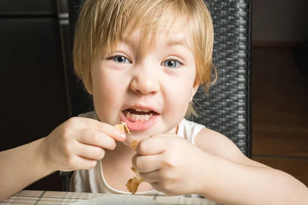 Beautiful baby with blue eyes eats delicious pancakes while sitting at kitchen table on chair. — Stock Photo, Image