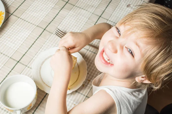 Beau bébé aux yeux bleus mange de délicieuses crêpes tout en étant assis à la table de cuisine sur une chaise. — Photo