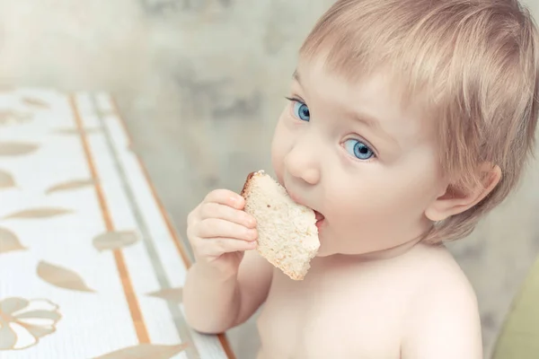 Красива дитина з блакитними очима сидить за кухонним столом і кусає шматочок білого хліба . — стокове фото