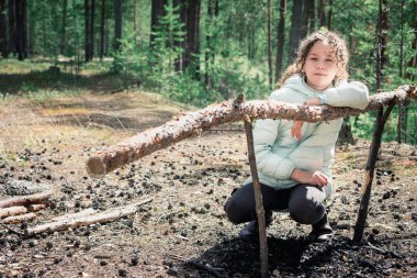 Kıvırcık ceketli kız orman yürüyüşünde yanan şenlik ateşinin yanında ve melon şapka takıyor.
