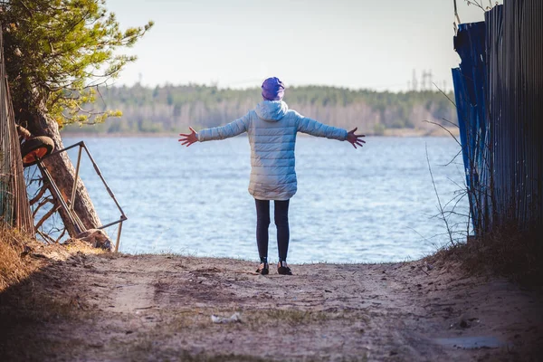 春の日に湖の海岸に立っている間 ダウンジャケットと帽子の女の子は側面に彼女の腕を伸ばし — ストック写真