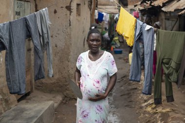 KAMPALA, UGANDA, JANUARY 26, 2020: Tanımlanamayan hamile kadın Katanga 'nın gecekondu mahallesinde asılı giysilerin yanından geçiyor, Kampala, Uganda. Yerel halk, kadın ve çocuklara düzgün bir sağlık hizmeti verilmeden burada kalmak zorunda..
