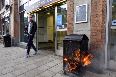 Stockholm, Sweden May 16, 2020 A pedestrian walks by a burning garbage can at the entrance to the Hornstull subway or Tunnelbana station. clipart