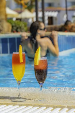 Yüzme havuzunun kenarında doğal meyve suyu ve buzlu iki kokteyl. Portakal dilimli portakal kokteyli ve kavun dilimli kırmızı. Arkaplanda canlı havuz suyu ve kız var.