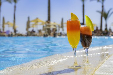 Yüzme havuzunun kenarında doğal meyve suyu ve buzlu iki kokteyl. Portakal dilimli portakal kokteyli ve kavun dilimli kırmızı. Arkaplanda canlı havuz suyu ve palmiye ağaçları var.