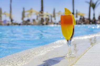 Yüzme havuzunun kenarında doğal meyve suyu ve buzlu iki kokteyl. Portakal dilimli portakal kokteyli ve kavun dilimli kırmızı. Arkaplanda canlı havuz suyu ve palmiye ağaçları var.