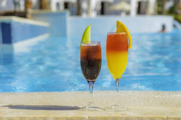 Two cocktails with natural juice and ice at the edge of the swimming pool. Orange cocktail with an orange slice and red with a melon slice with backdrop of vibrant pool water