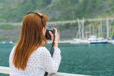 Kameralı güzel bir kızıl saçlı kız marinanın gün ışığında fotoğrafını çekiyor. Kanarya adalarında seyahat fotoğrafı çeken bir kadın. Arka planda yumuşak odaklı yatlar.