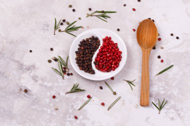 Beyaz bir tabakta kırmızı ve siyah bezelye biberi. Kasenin yanında tahta bir kaşık ve biberiye yaprakları var..