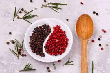 Beyaz bir tabakta kırmızı ve siyah bezelye biberi. Kasenin yanında ahşap kaşık ve biberiye yaprakları var. Yakın plan. Makro çekim
