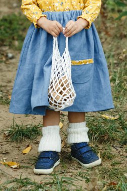 Çocuk fındıklı bir ağ çanta tutuyor. Mavi elbiseli, mavi ayakkabılı, fındıklı örgü.