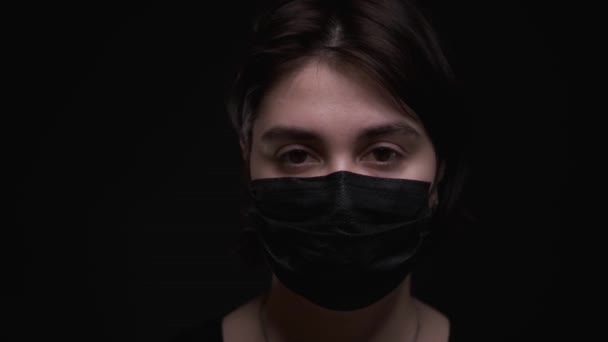 Caucásico chica en negro máscara médica, mirando a la cámara, fondo negro — Vídeos de Stock