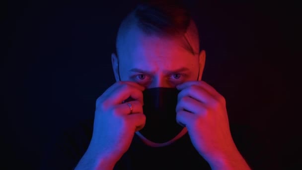 Blanke man zet op zwart medisch masker, rood en blauw neon licht op het gezicht — Stockvideo