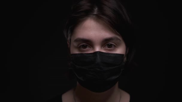 Caucasian girl in black medical mask,coughing at camera,black background — Stock Video