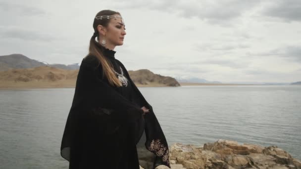 Жінка в чорній сукні хіджаб, сая, стоїть на краю скелі біля озера, дивиться попереду — стокове відео