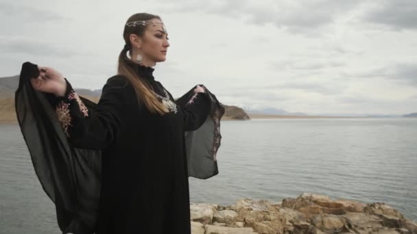 Mujer en vestido negro hijab, abaya, se encuentra en el borde del acantilado cerca del lago, mira hacia el futuro — Vídeos de Stock