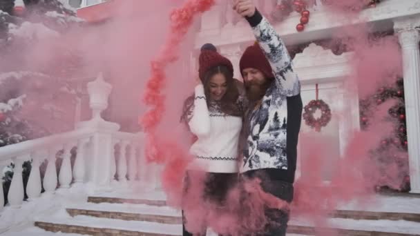 คู่รักหนุ่มสาวในวันคริสต์มาส ปีใหม่เล่นระเบิดควันสีชมพูใกล้บ้านตกแต่ง — วีดีโอสต็อก