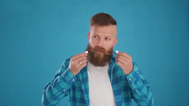 Homme caucasien insère écouteurs ses oreilles, écoute de la musique, sur fond bleu — Video