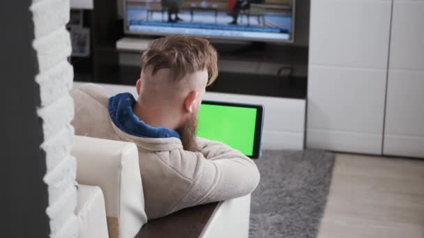 Hombre trabaja en casa, mira la pantalla de la tableta, cuarentena frente a la televisión con noticias — Vídeos de Stock