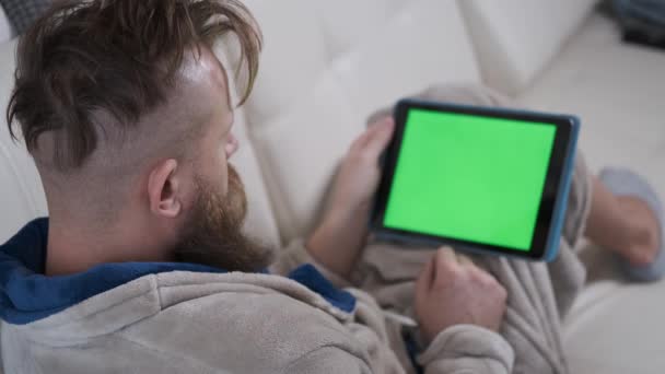 Hombre en la avena de casa se encuentra en el sofá y voltea a través de la pantalla de una tableta cromakey, trabajando en casa — Vídeos de Stock