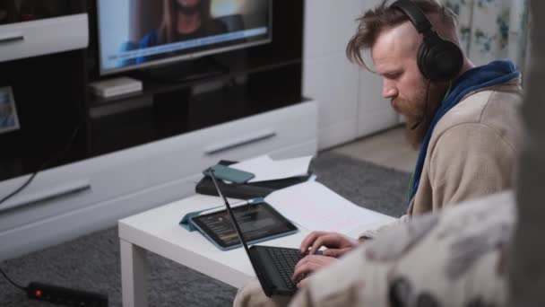 Uomo lavora a casa in grandi cuffie, digitando sul computer portatile, e guardando le notizie — Video Stock