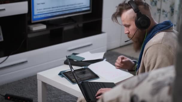 Man werkt thuis in grote koptelefoons, typen op laptop, en kijken naar nieuws — Stockvideo