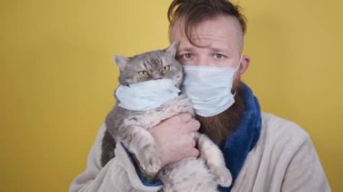 Maskeli bir adam sarı arka planda maskeli bir kedi tutuyor.