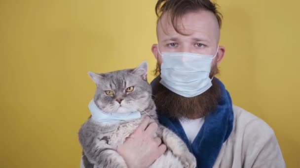 Man met medisch masker houdt gemaskerde kat in zijn armen geïsoleerd op gele achtergrond — Stockvideo