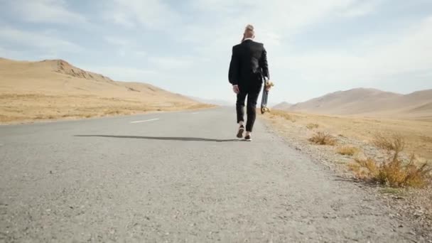 Uomo in completo nero cammina lungo solitarie strade desertiche in montagna con longboard — Video Stock