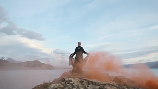 Frau in schwarzem Kleid steht malerisch am Rand der Klippe, signalisiert Feuer, roter Rauch — Stockvideo