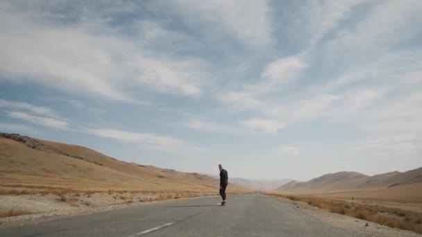 Homme en costume noir marche le long de la route déserte solitaire dans les montagnes avec planche à roulettes — Video
