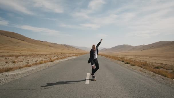 Hombre en traje negro camina a lo largo del solitario camino del desierto en las montañas con monopatín — Vídeos de Stock