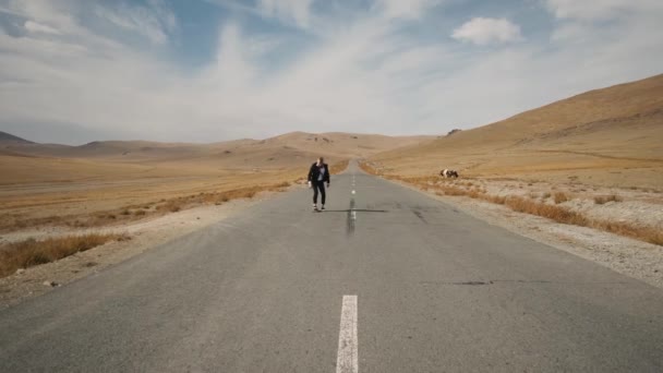 Muž v černém obleku procházky podél osamělé pouštní silnici v horách s skateboard — Stock video