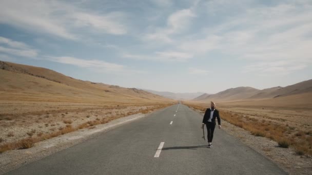 Homem de terno preto caminha ao longo da estrada deserto solitário em montanhas com skate — Vídeo de Stock