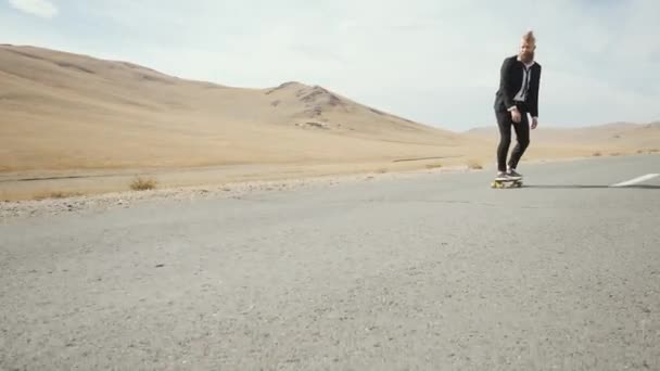 Man in zwart pak loopt langs eenzame woestijn weg in de bergen met skateboard — Stockvideo