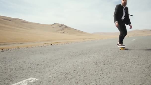 Homem de terno preto caminha ao longo da estrada deserto solitário em montanhas com skate — Vídeo de Stock