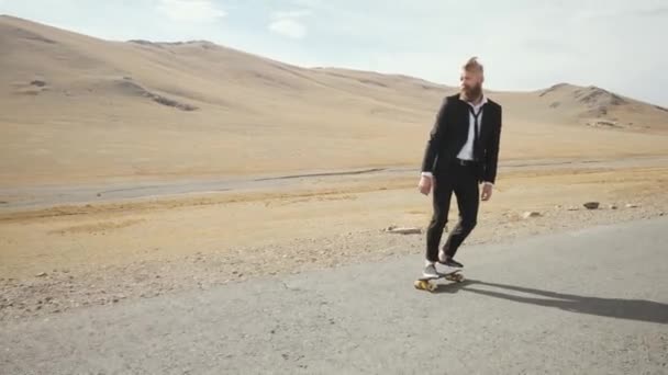 Man in zwart pak loopt langs eenzame woestijn weg in de bergen met skateboard — Stockvideo