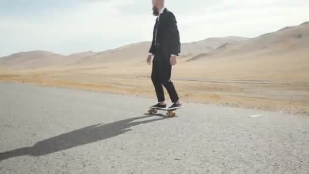 Férfi fekete öltönyben sétál magányos sivatagi úton a hegyekben gördeszkával — Stock videók
