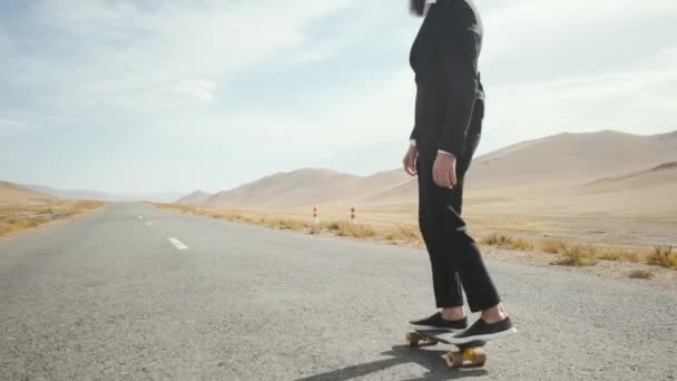 Man in zwart pak loopt langs eenzame woestijn weg in de bergen met skateboard — Stockvideo