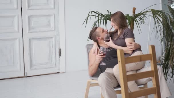 Молодая счастливая супружеская пара обнимает друг друга сидя дома и попивая красное вино — стоковое видео