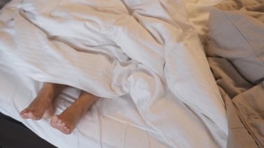 Yatak odasındaki yorganın altından çıkan genç bir kadının ayaklarına yakın çekim. Çıplak ayaklar