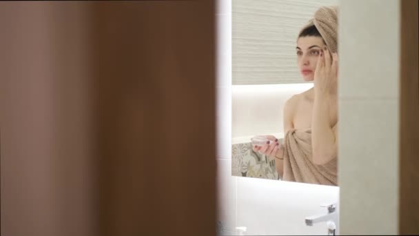 Jeune femme appliquant crème visage à la maison dans la salle de bain, en face du miroir — Video