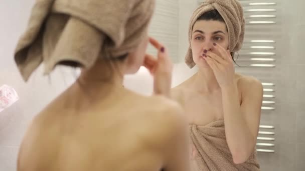 Молодая женщина наносит крем для лица дома в ванной комнате, перед зеркалом — стоковое видео