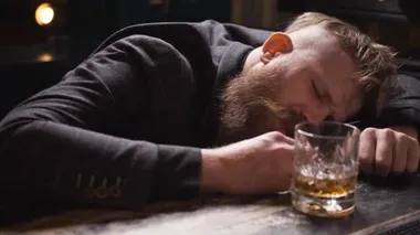 Sarhoş adam bir bardak viskiyle uyuya kaldı, kötü ve üzgün görünüyor.