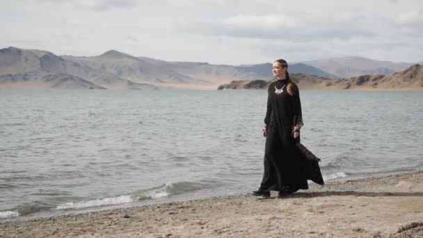 Hermosa chica camina a lo largo de las orillas de un río, cerca de grandes montañas, vestido de luto — Vídeos de Stock