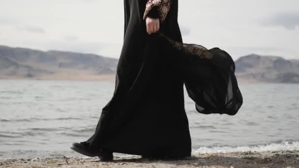 Chica caucásica camina a lo largo de la orilla del lago, vestido de luto negro, mujer camina sobre rocas — Vídeo de stock