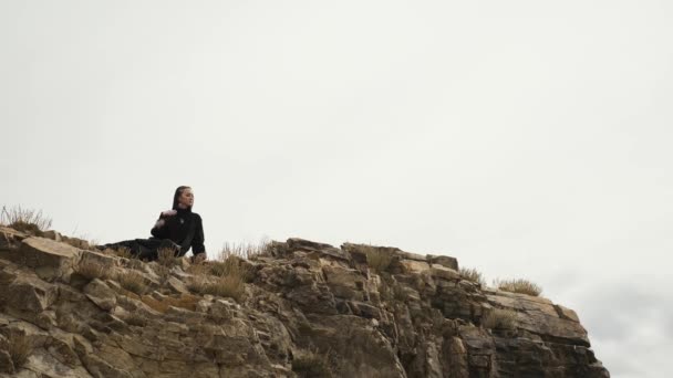 Мусульманка девушка сидит на краю скалы на вершине горы, большое озеро — стоковое видео