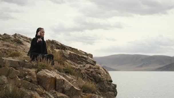 Siyah elbiseli kadın dağın tepesindeki uçurumun kenarında oturuyor. Büyük göl. — Stok video