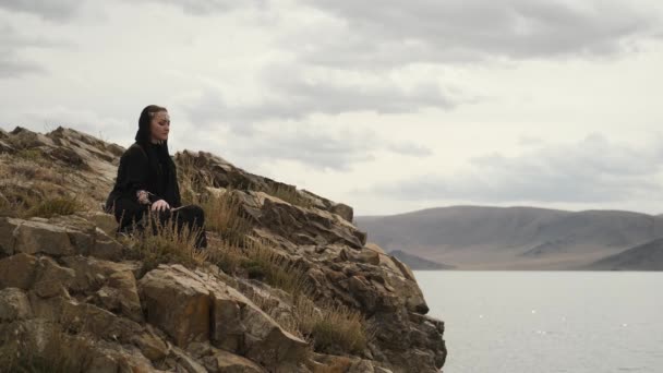 Kvinna i svart klänning sitter på kanten av klippan på toppen av berget.stor sjö — Stockvideo
