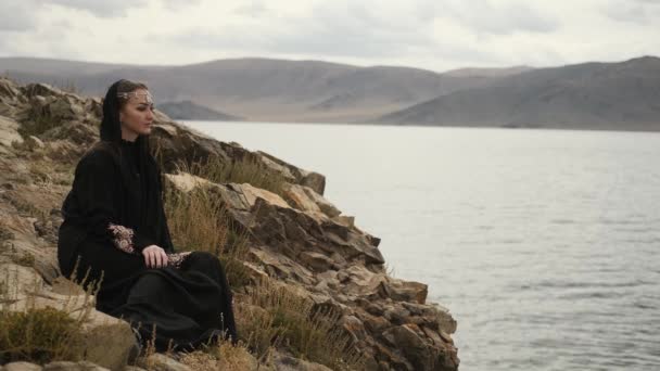 Siyah elbiseli kadın, dağın tepesinde uçurumun kenarında oturuyor. — Stok video